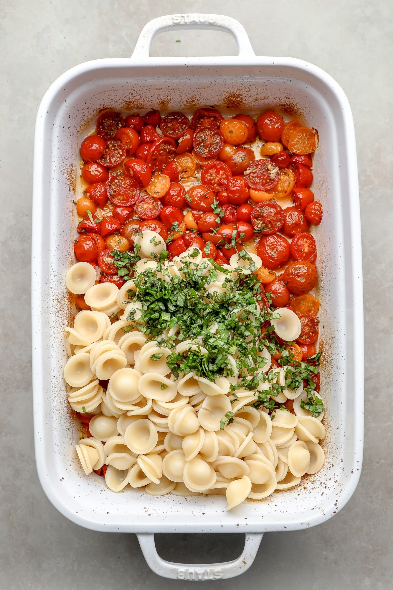 Macarrão cozido e ervas frescas em cima de tomates assados ​​em uma assadeira branca.